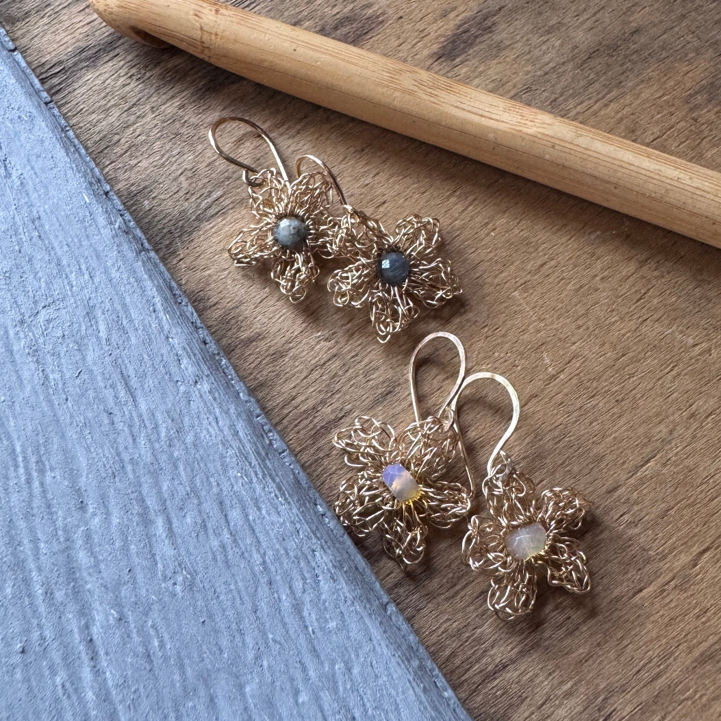 A pair of handmade gold wire flower dangle earrings with gray and iridescent opal-like center stones, displayed on a wooden surface next to a blue painted wood block.