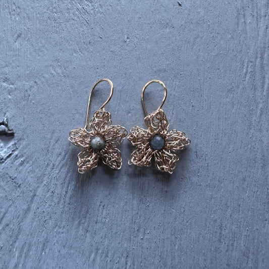 A pair of handmade gold-tone flower dangle earrings with small gray-blue stone centers, resting on a textured blue-gray wooden surface.