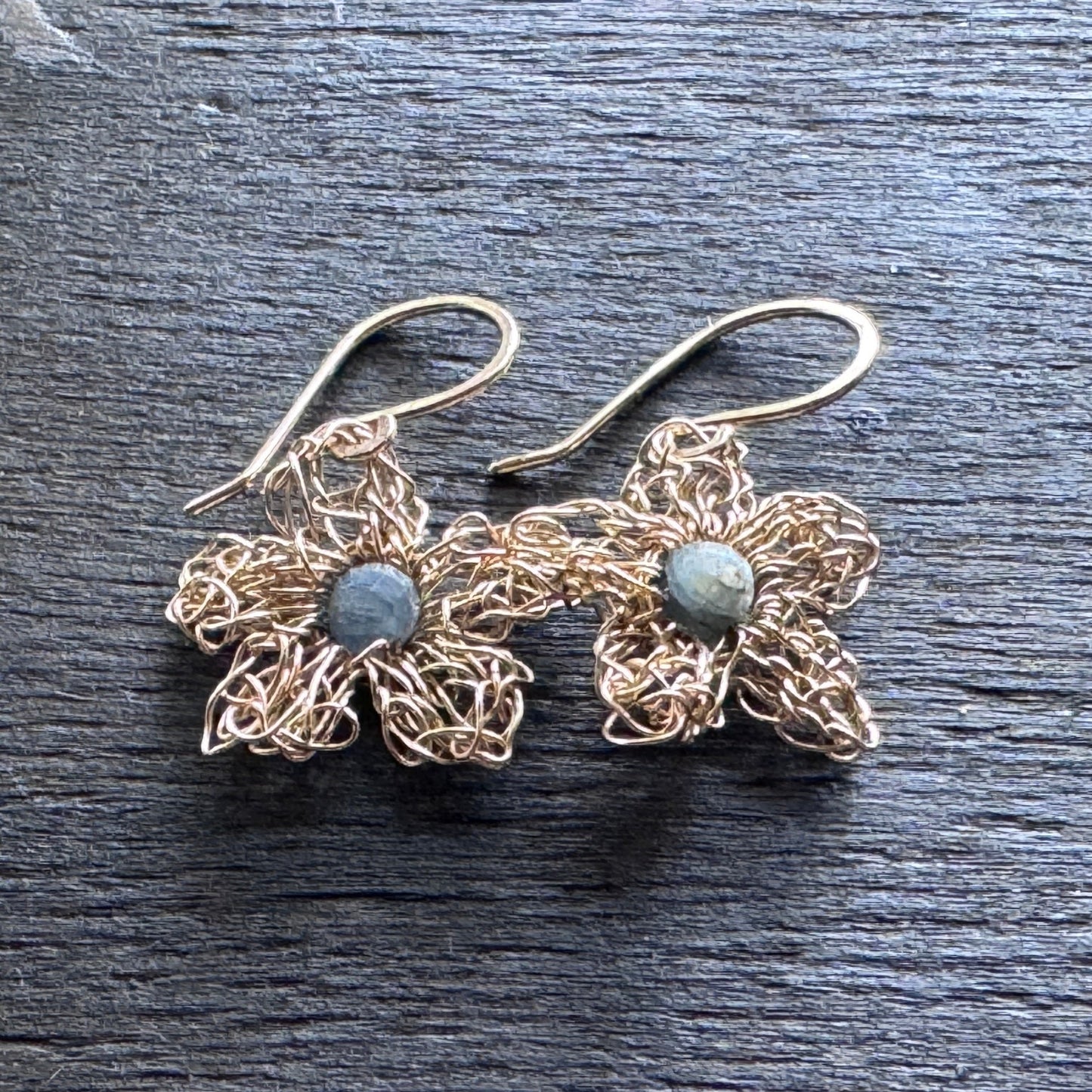 A pair of handmade wire-wrapped flower earrings with gold-toned petals, a small round gray stone in the center of each flower, and gold fishhook ear wires, displayed on a dark textured slate surface.