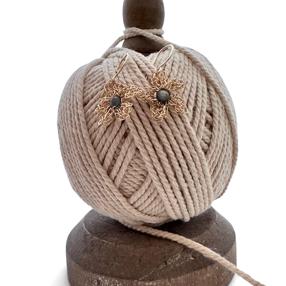 A pair of handmade flower-shaped earrings with gold-tone wire petals and dark round beads, displayed on top of a beige ball of yarn wound around a wooden spool against a white background.