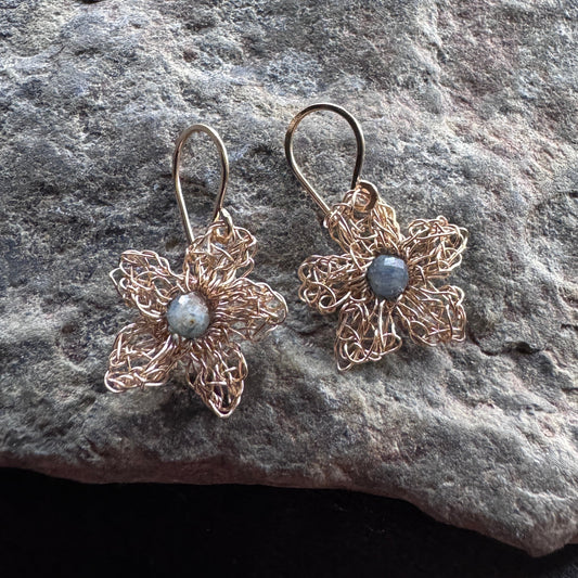 A pair of handmade wire-wrapped flower drop earrings in a coppery rose-gold tone, featuring a small round gray stone in the center of each flower, displayed on a textured gray rock.