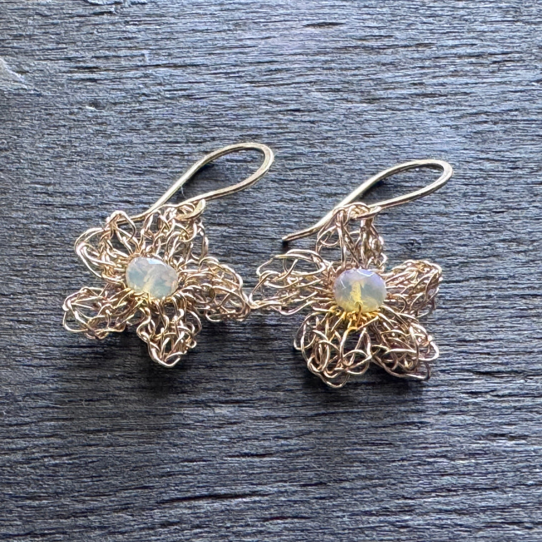 A pair of handmade flower-shaped dangle earrings made of gold-toned wire with round iridescent opal gemstones in the center, displayed on a dark textured wooden surface.
