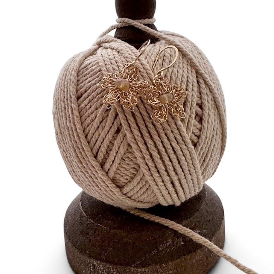 A pair of gold-toned flower-shaped earrings with small clear stones, displayed on a ball of beige yarn wrapped around a dark wooden stand against a white background.