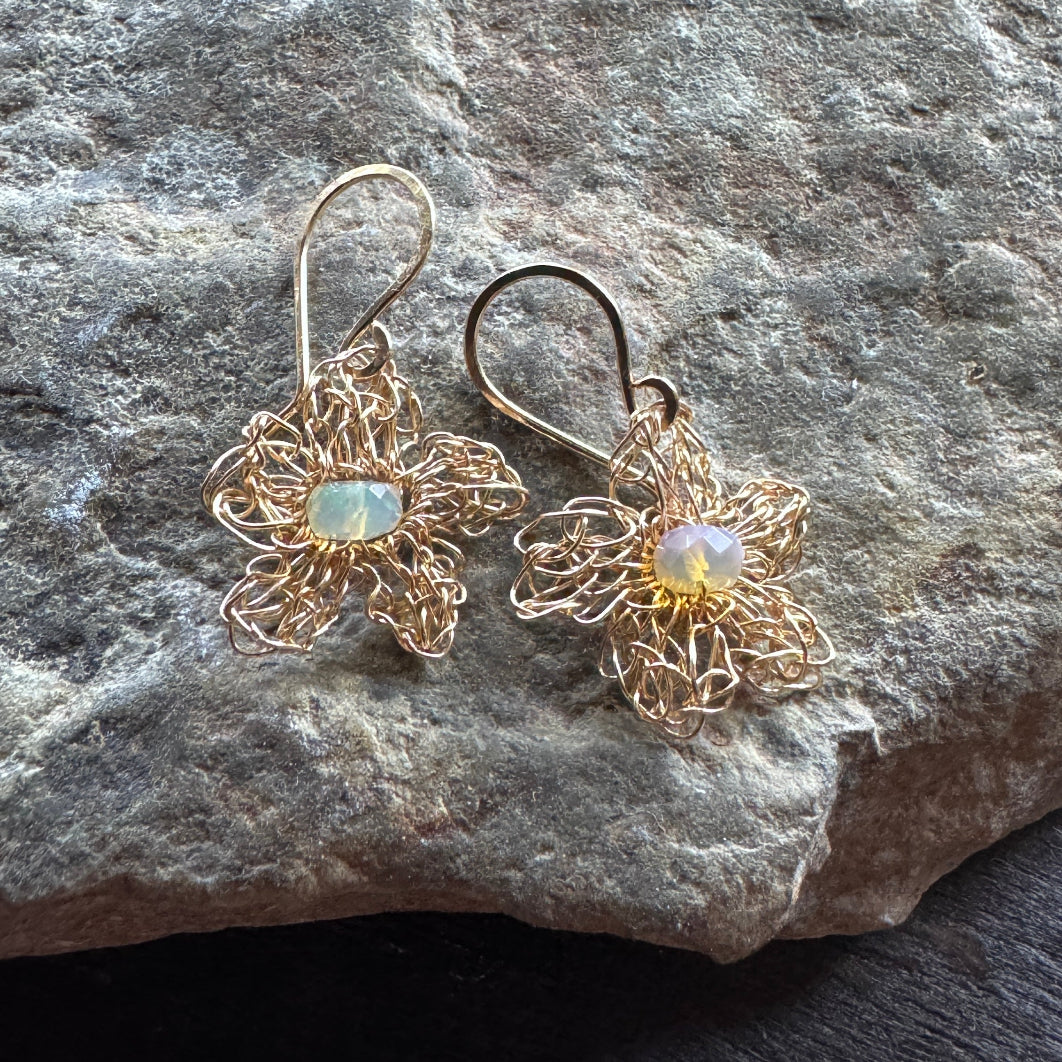 A pair of handmade wire-wrapped flower dangle earrings in a gold-tone finish, featuring iridescent opal gemstones at the center, displayed on a textured gray stone surface.