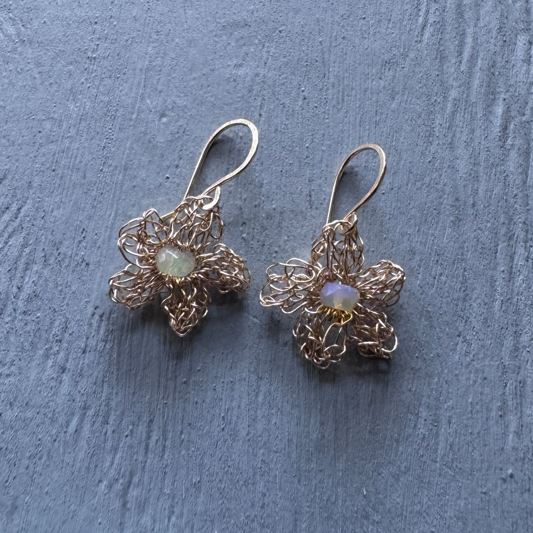 A pair of handmade flower-shaped dangle earrings made of gold-toned wire with iridescent opal-like centers, resting on a textured gray surface.