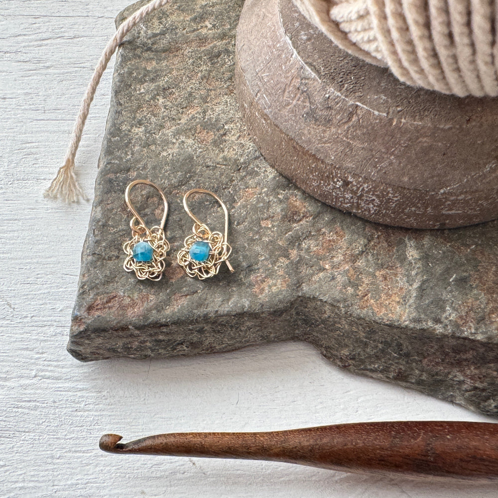 Gold earrings with blue stones on a stone surface next to a wooden crochet hook.