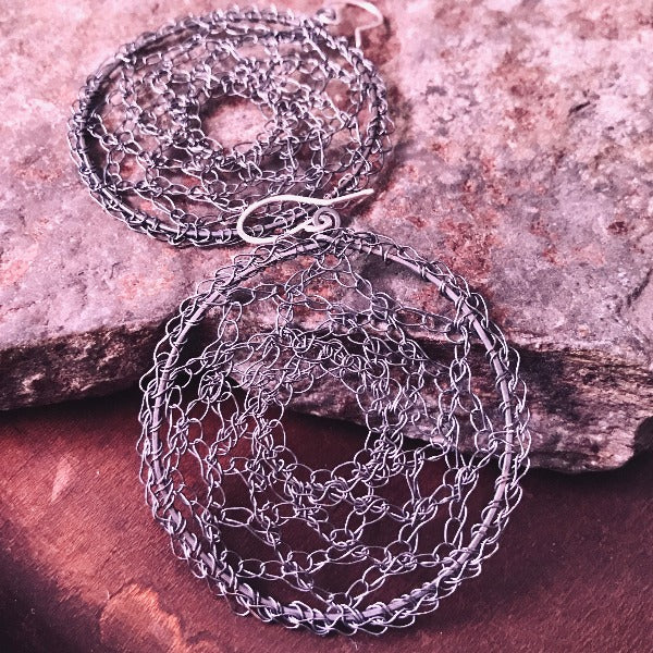 A pair of crocheted silver hoop earrings with an intricate lace pattern, displayed against a textured background.